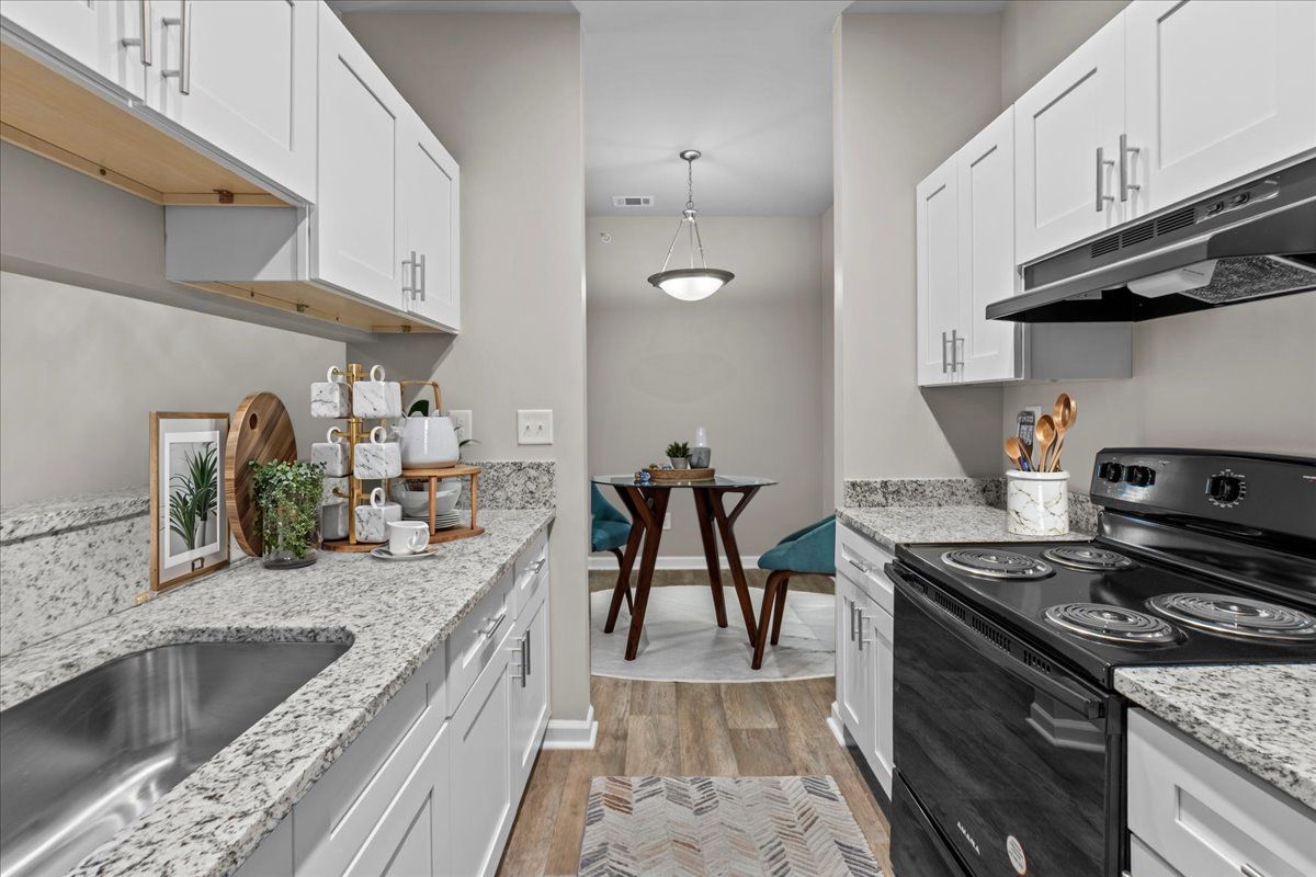 A modern galley kitchen with white cabinets, speckled countertops, a black stove, and a dining area in the background.