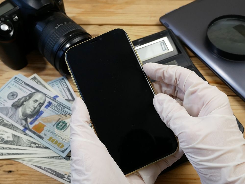 Gloved hands holding a black smartphone beside cash, camera, and laptop on a desk