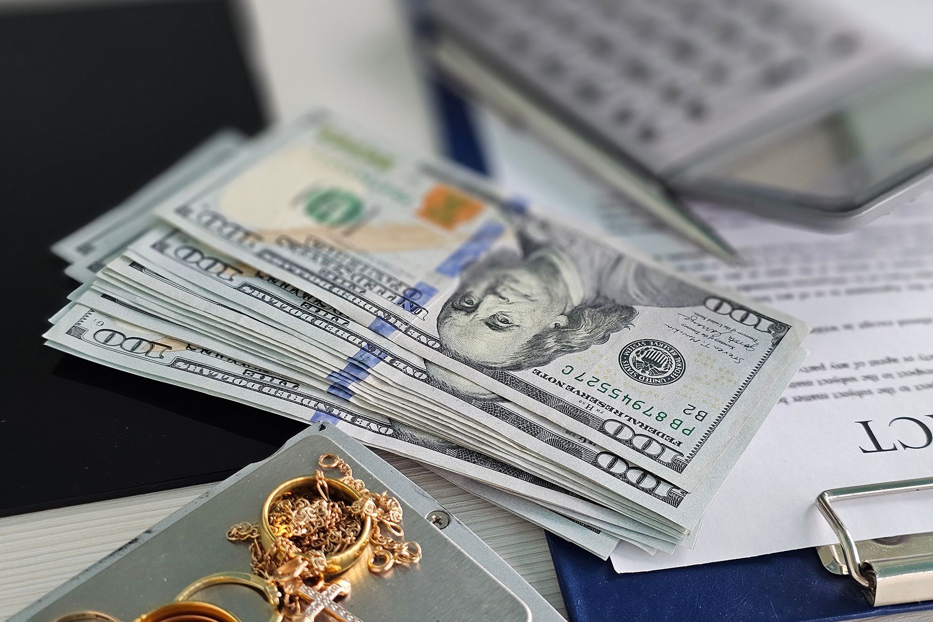 Stack of US dollar bills beside a laptop, clipboard, and gold necklace on a desk.