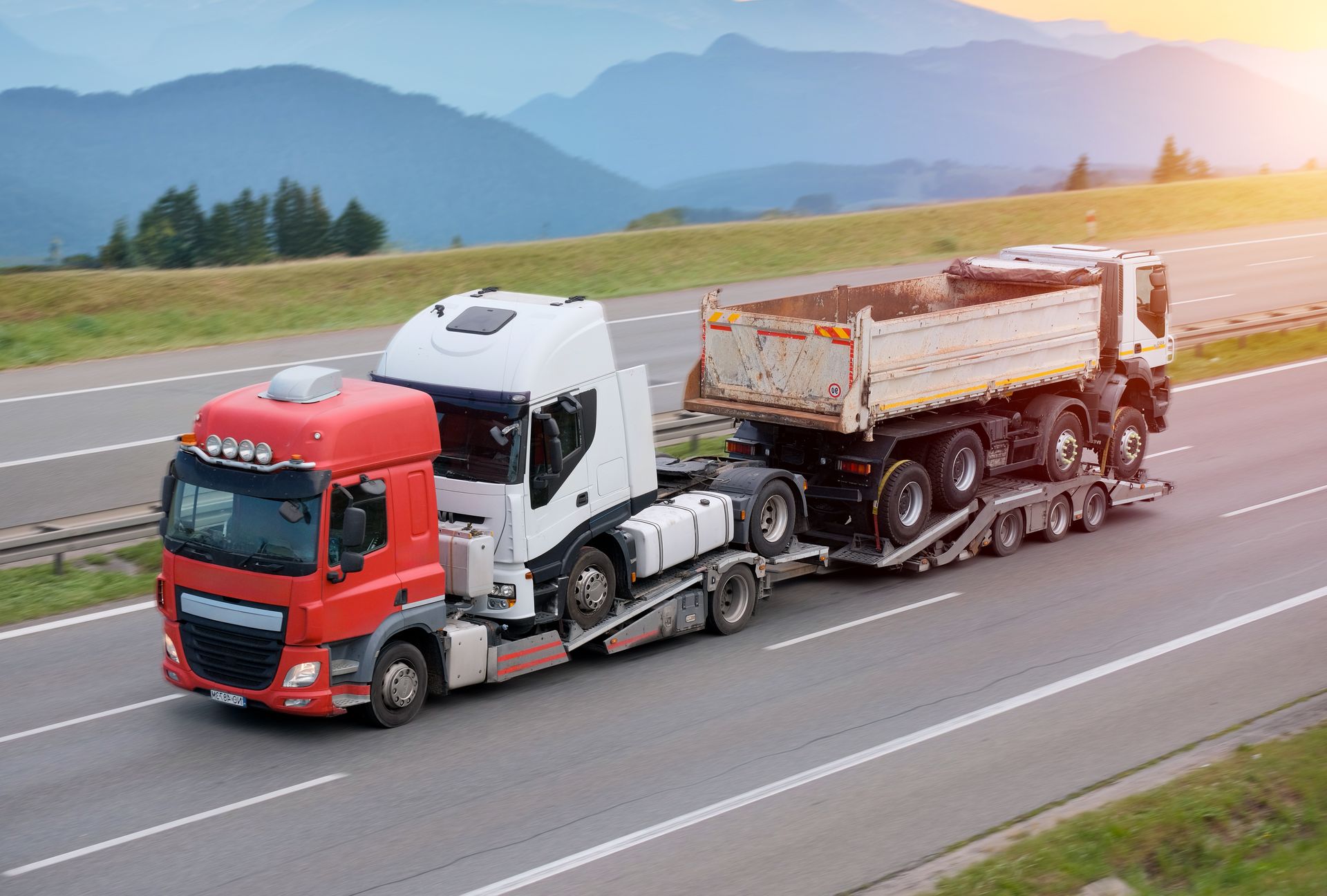 Truck hauling two large vehicles on a highway with mountains in the background.