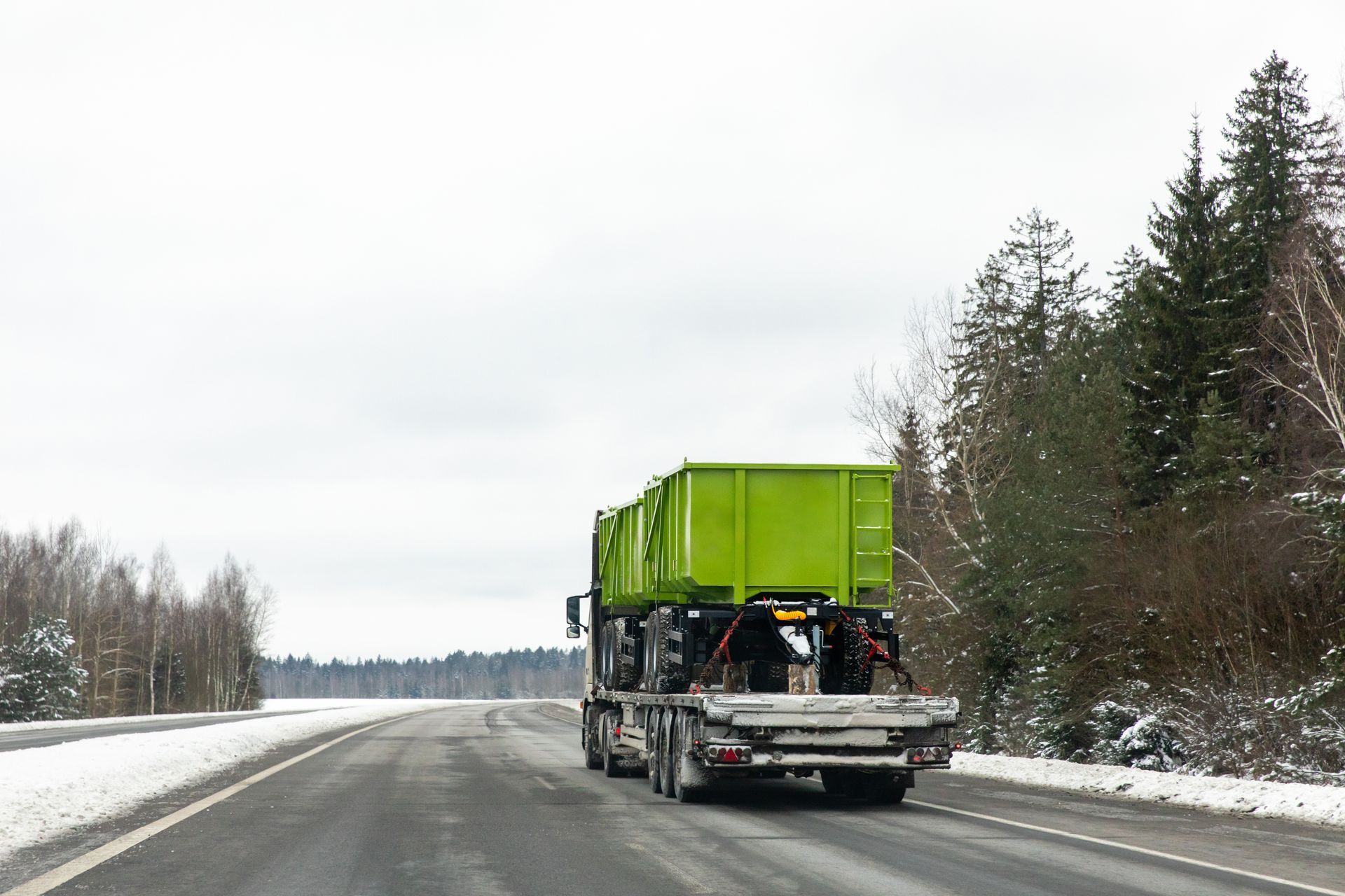 Flatbed semi truck hauling a large green industrial container down a snowy highway.