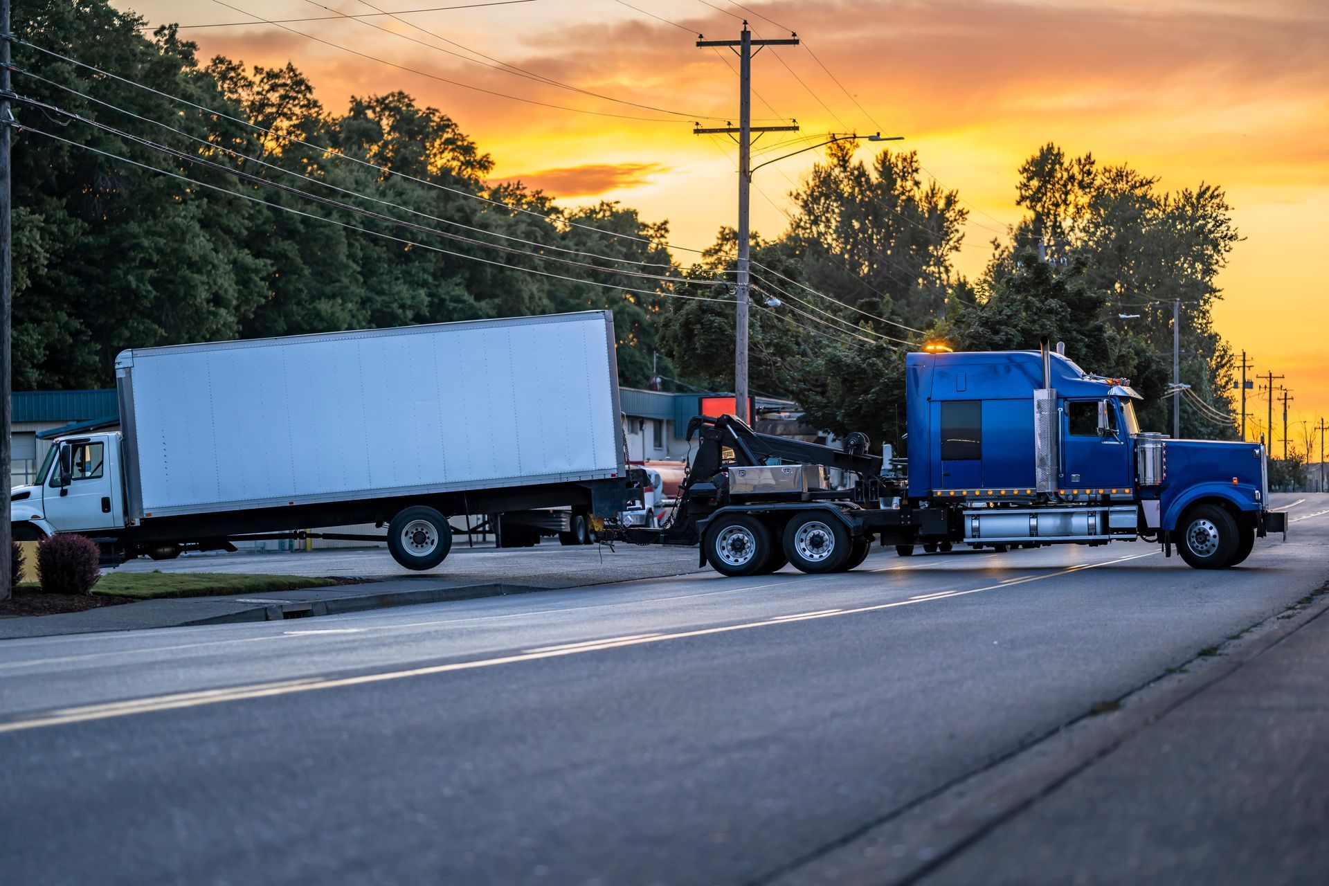 A heavy-duty blue tow truck hauling a broken semi-truck with a box trailer at sunset. A heavy-duty blue tow truck hauling a broken semi-truck with a box trailer at sunset.