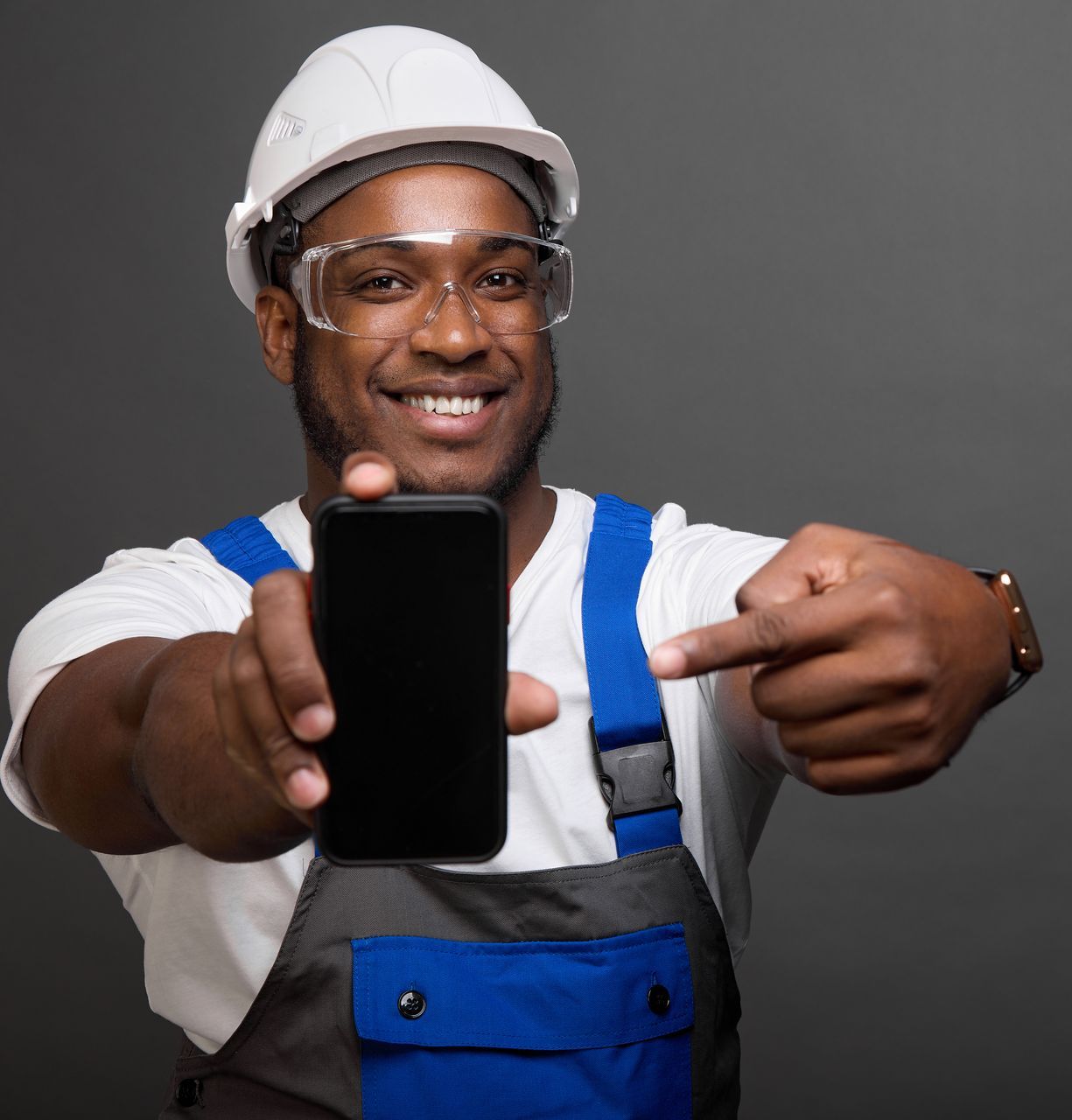 A man wearing a hard hat and goggles is pointing at a cell phone