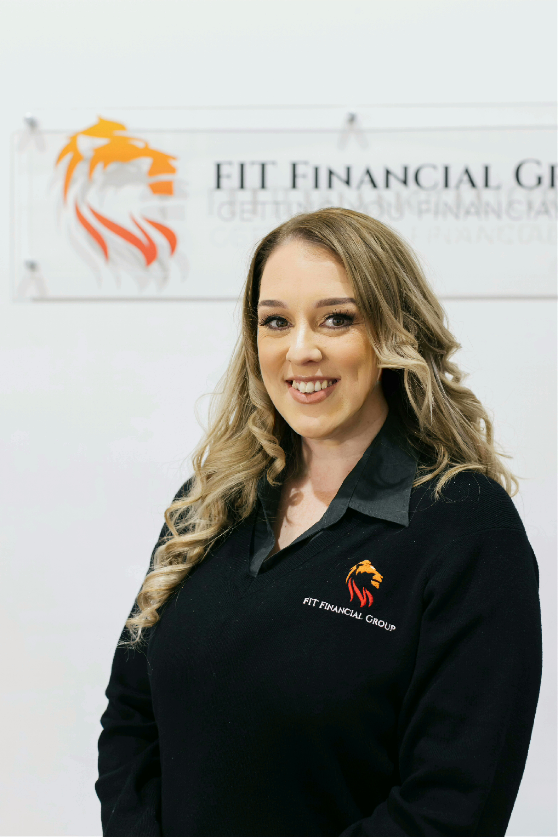 Woman in black shirt with logo, smiling in front of a sign that also has the same logo — Fit Financial Group in Leumeah, NSW