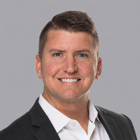 A man in a suit and tie is smiling in front of a brick wall.
