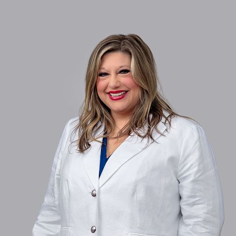 A woman in a blue shirt and black jacket is smiling in front of a brick wall.