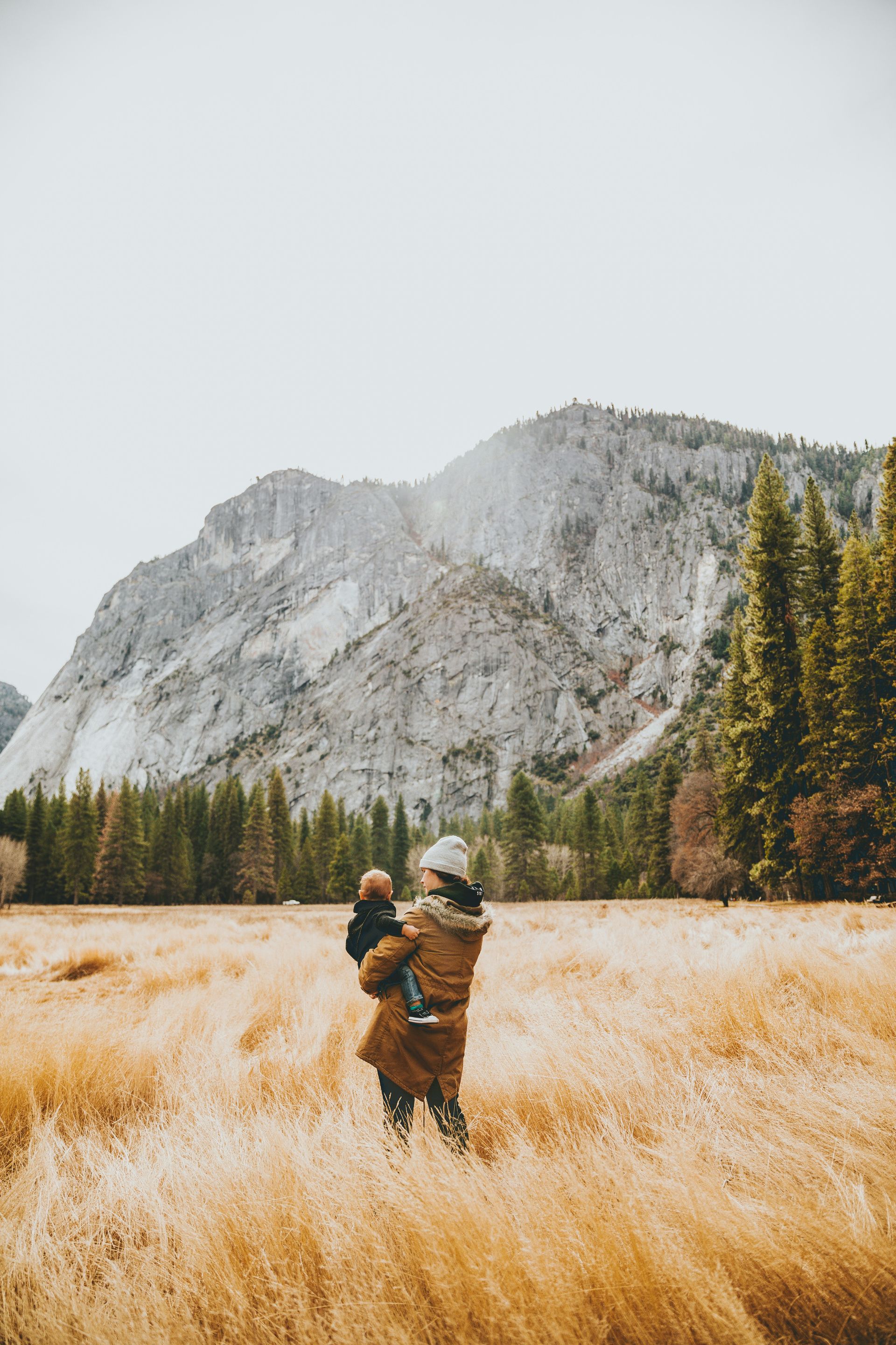 A woman is holding a baby in a field with mountains in the background.