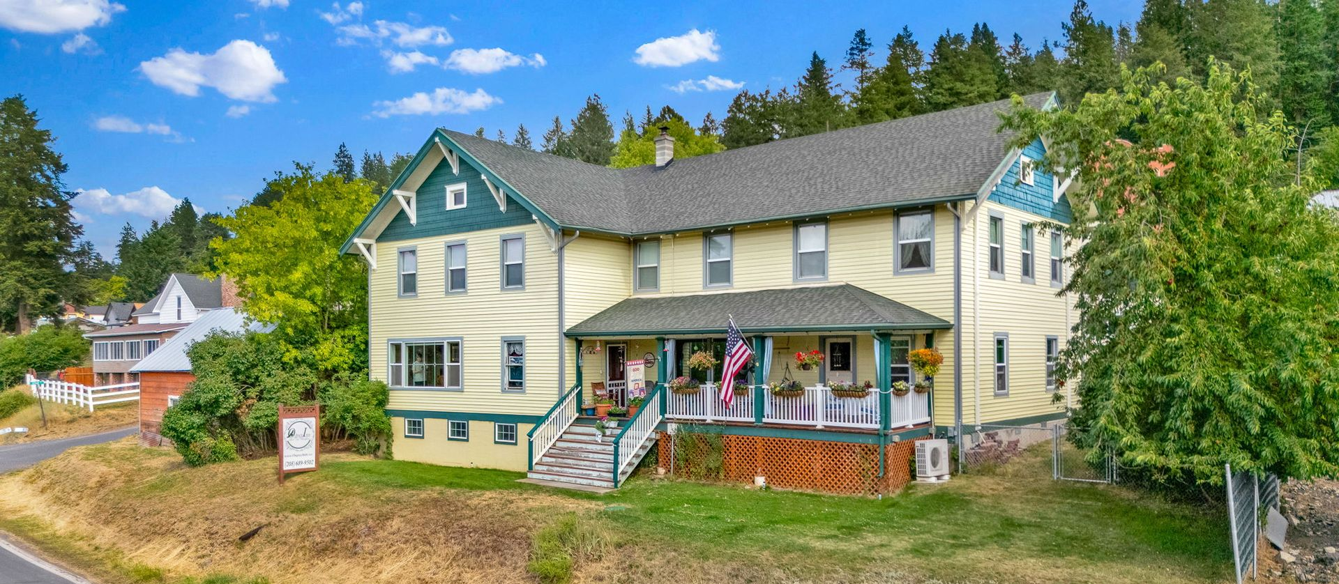 Yellow house with green trim and a front porch on a sunny hillside, surrounded by trees and blue sky