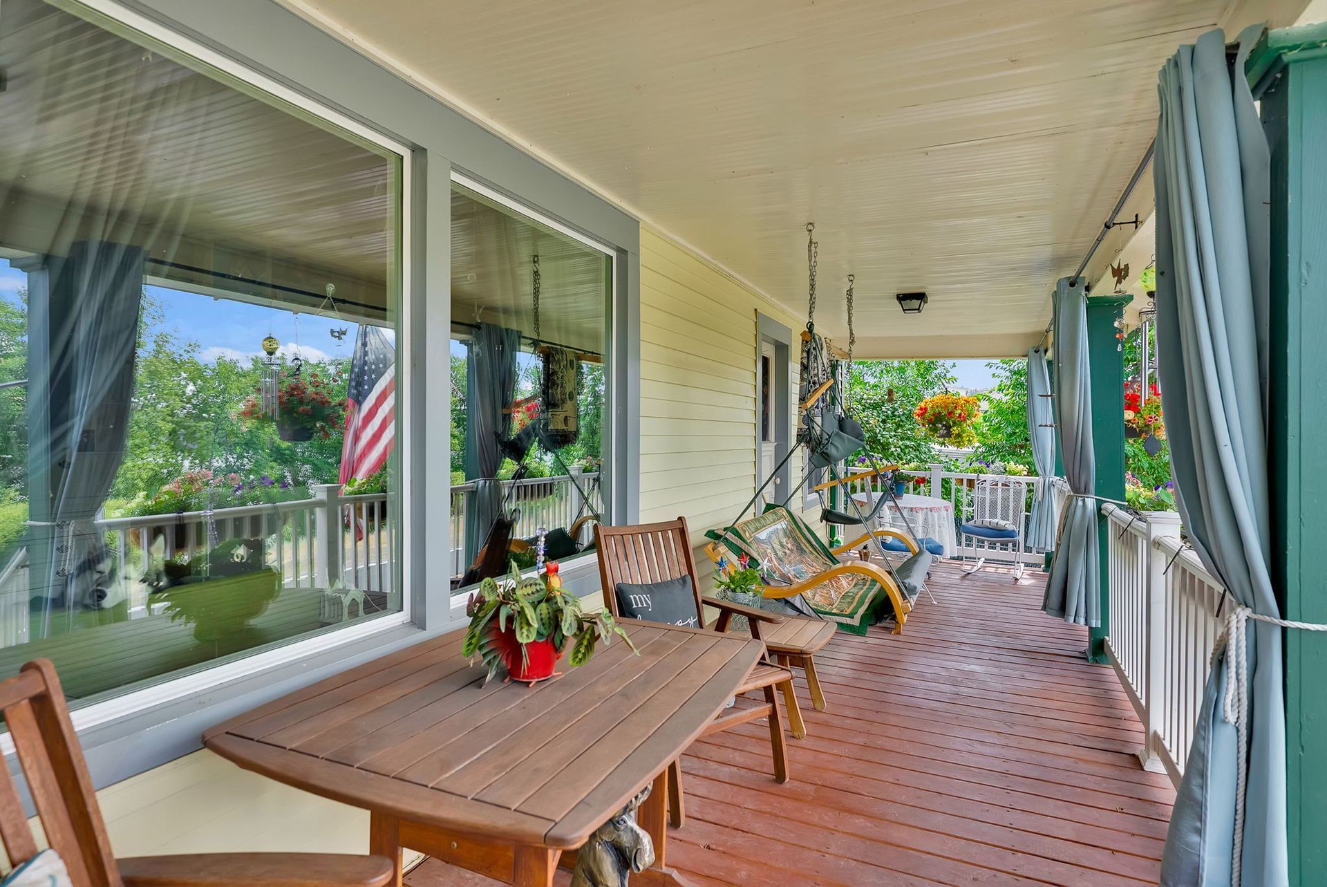 Covered porch with wooden table, chairs, potted flowers, and a hanging swing overlooking a garden.