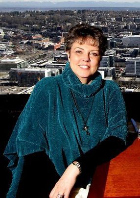 Woman in a teal shawl seated by a window, with a cityscape visible in the background.