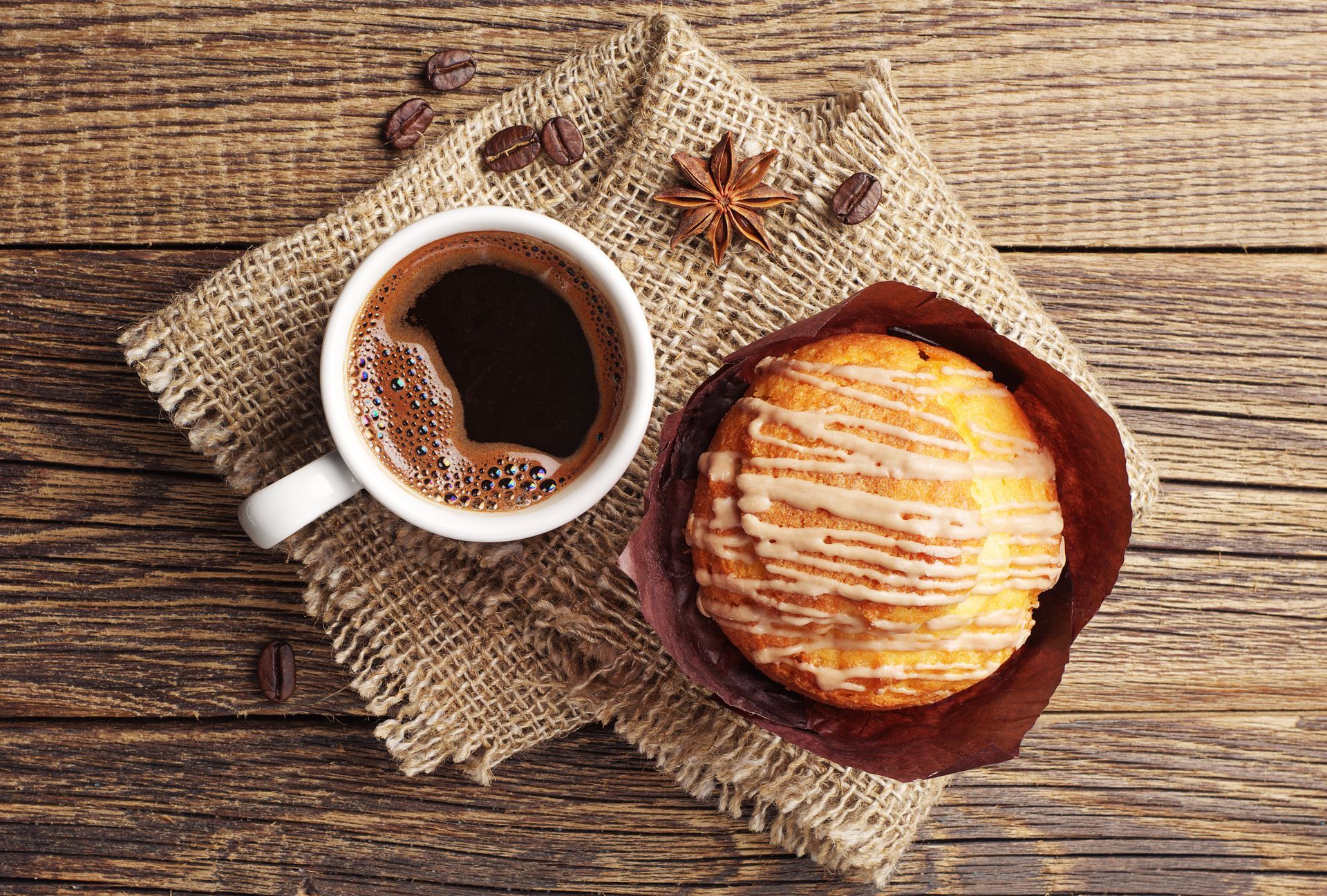 Cup of coffee and a glazed pastry on burlap cloth, with star anise on a rustic wooden table