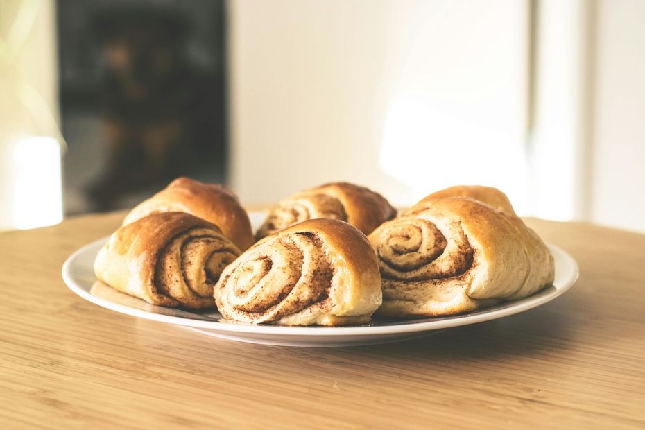 A plate of fresh, golden-brown cinnamon rolls rests on a wooden table in a brightly lit room.