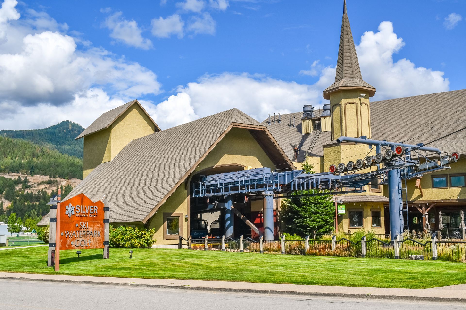 A large, tan, multi-gabled building with a turret, featuring ski lift machinery and a wooden sign on a lush green lawn.