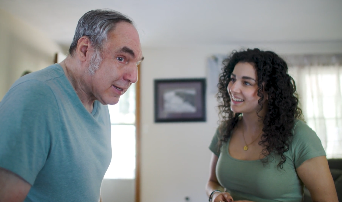 A man in a teal shirt looks toward a smiling woman with dark curly hair in a green shirt inside a home.