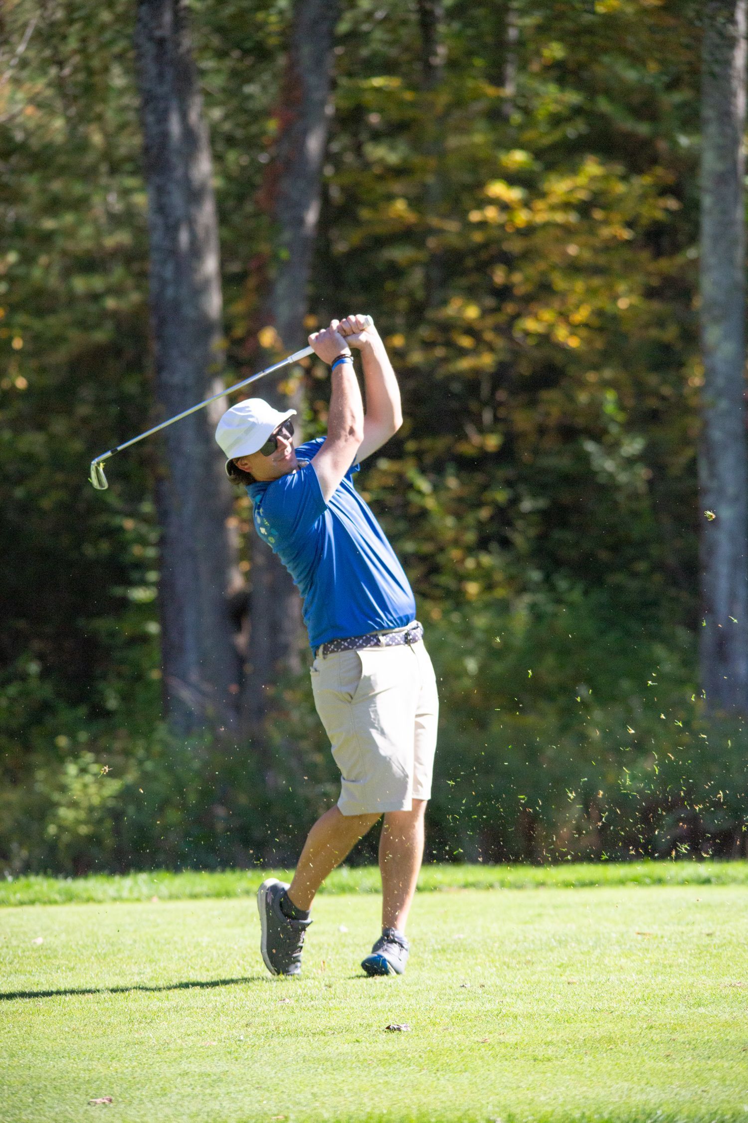 Golfer in blue shirt and khaki shorts swings a club on a golf course.