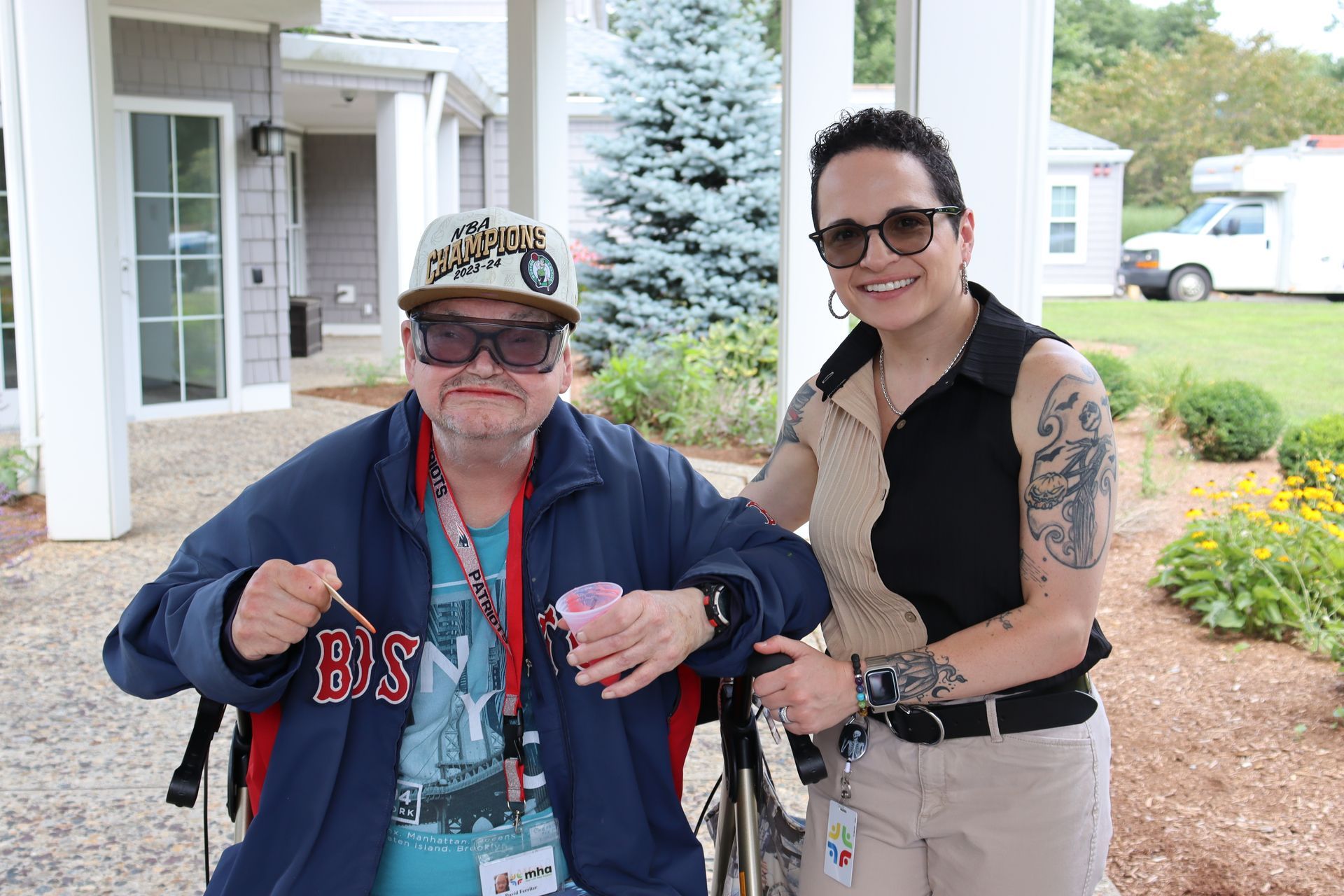 A person in a wheelchair wearing a baseball cap and dark glasses stands next to a woman with arm tattoos outside a building.