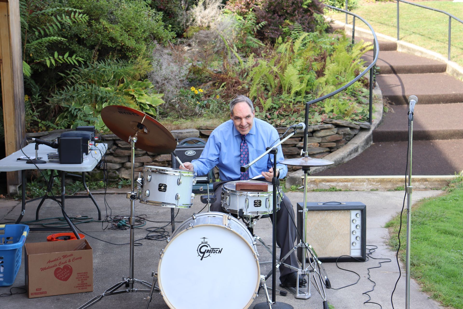 A person plays a Gretsch drum kit outdoors on a stone patio with greenery in the background.