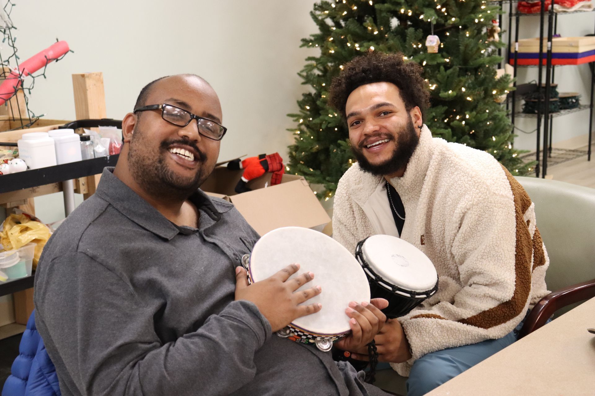 Two men are sitting at a table playing drums in front of a Christmas tree.