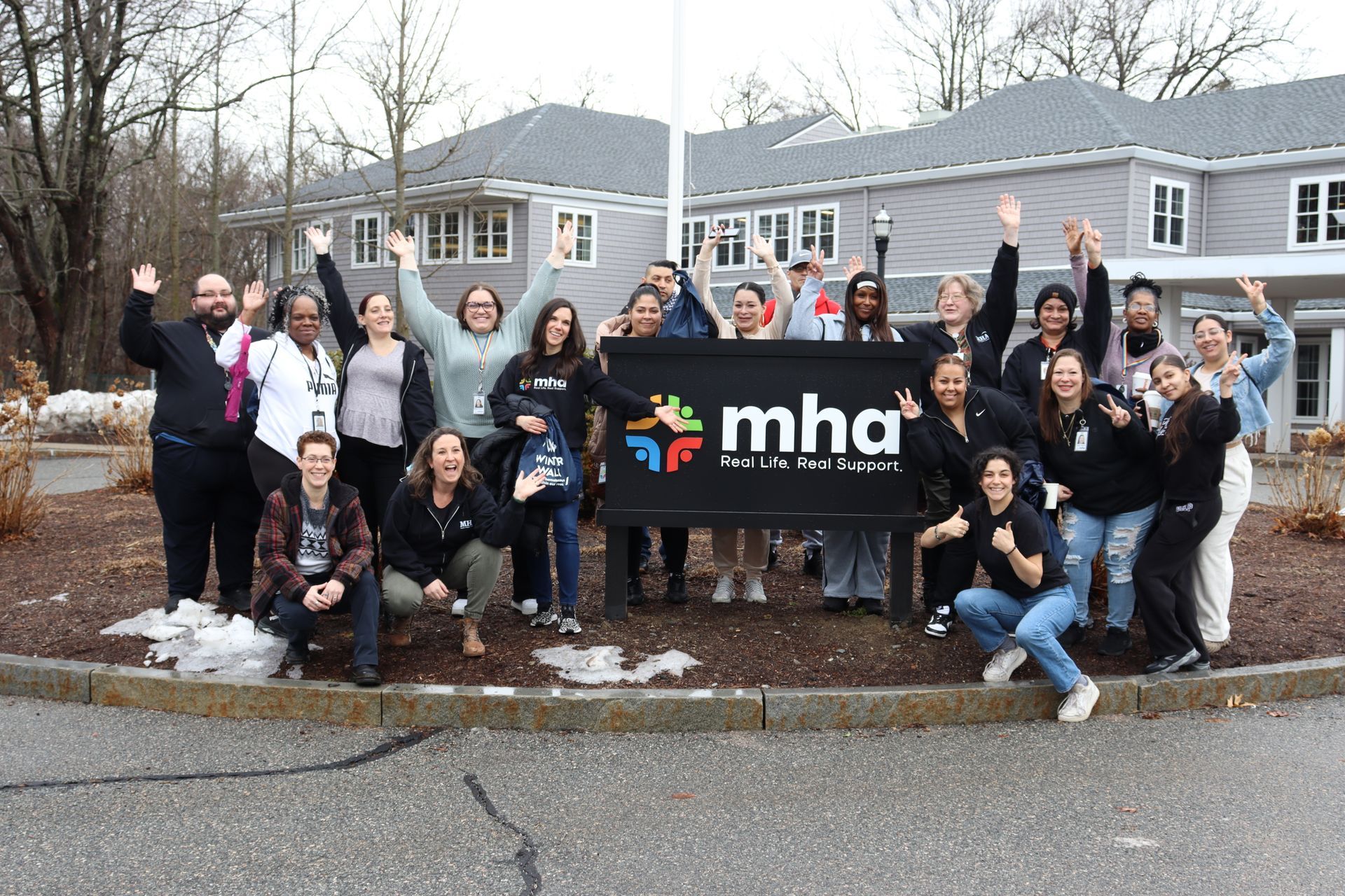 A group of people smiles and waves behind a sign for MHA in front of a gray, two-story building on a sunny day.