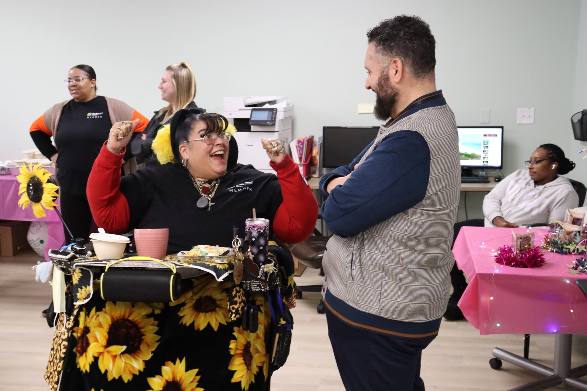 A joyful person in a sunflower-print wheelchair interacts with a standing person in a grey vest in an office setting.