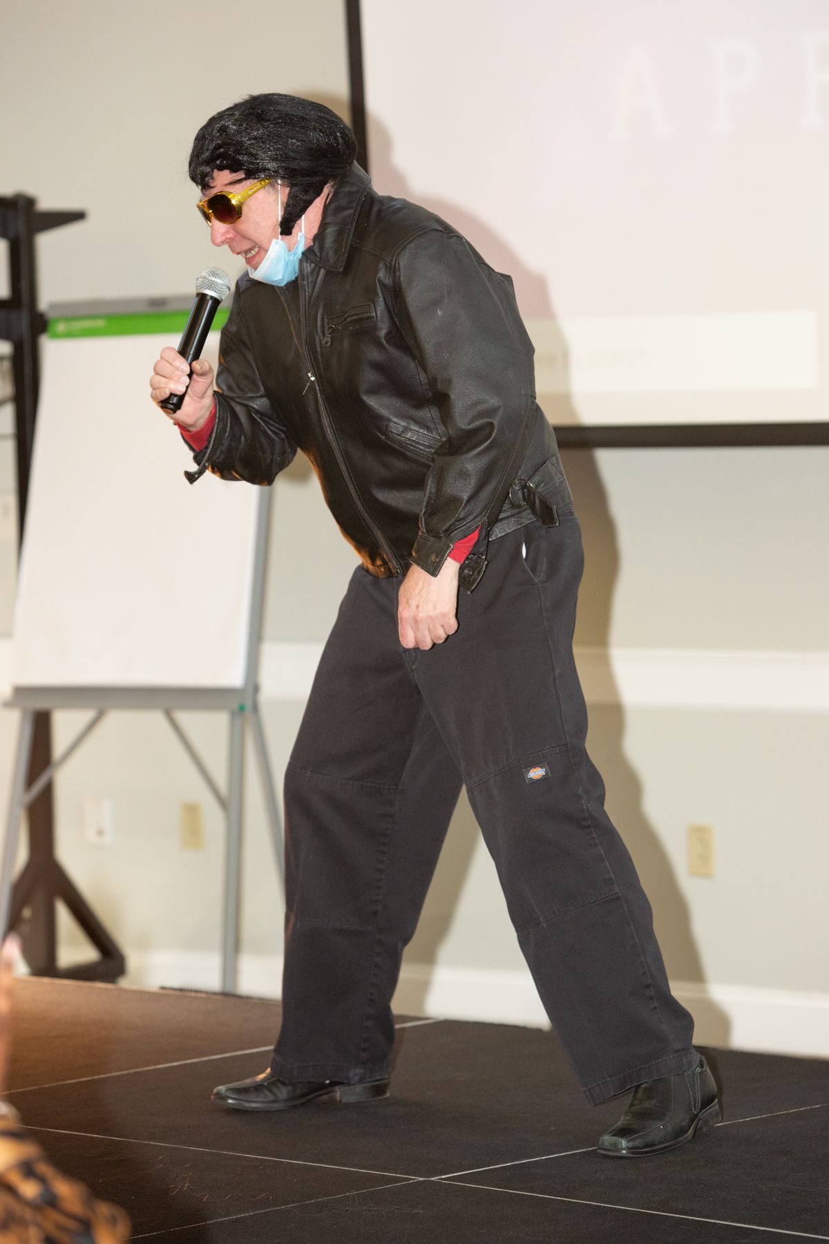 A person in an Elvis costume with a black jacket and sunglasses holds a microphone, performing on a stage.