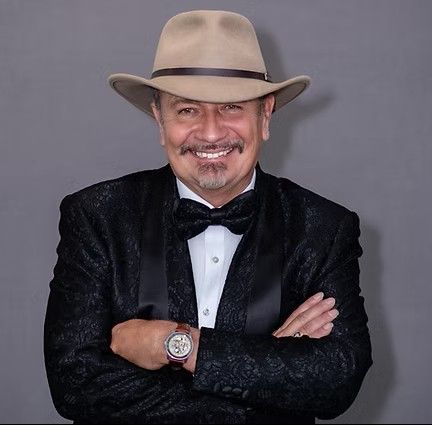 Man wearing a cowboy hat, tuxedo, and bow tie, arms crossed, smiling.