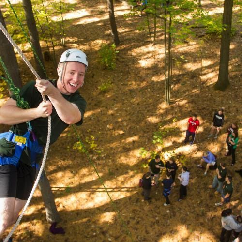 Man on a rope climbing course, smiling. Surrounded by group of people in a wooded area.