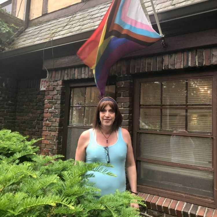 A woman stands in front of a building with a rainbow flag hanging from the roof