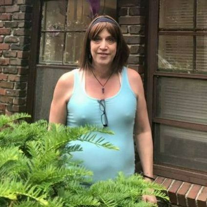 A woman in a blue tank top and glasses is standing in front of a brick building.