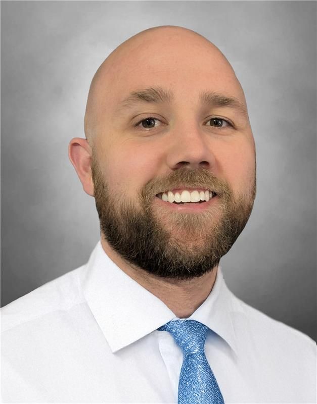 Professional headshot of a bald person with a beard, smiling, wearing a white shirt and blue patterned tie against gray.