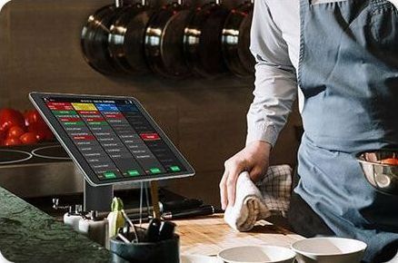 A man in an apron is standing in front of a tablet in a kitchen.