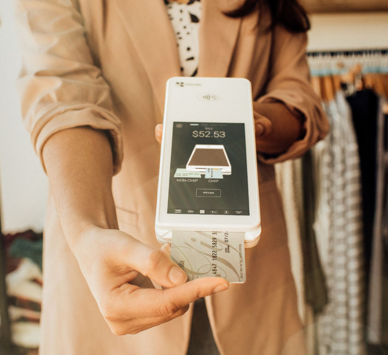 A woman in a tan jacket is holding a smart phone and a credit card