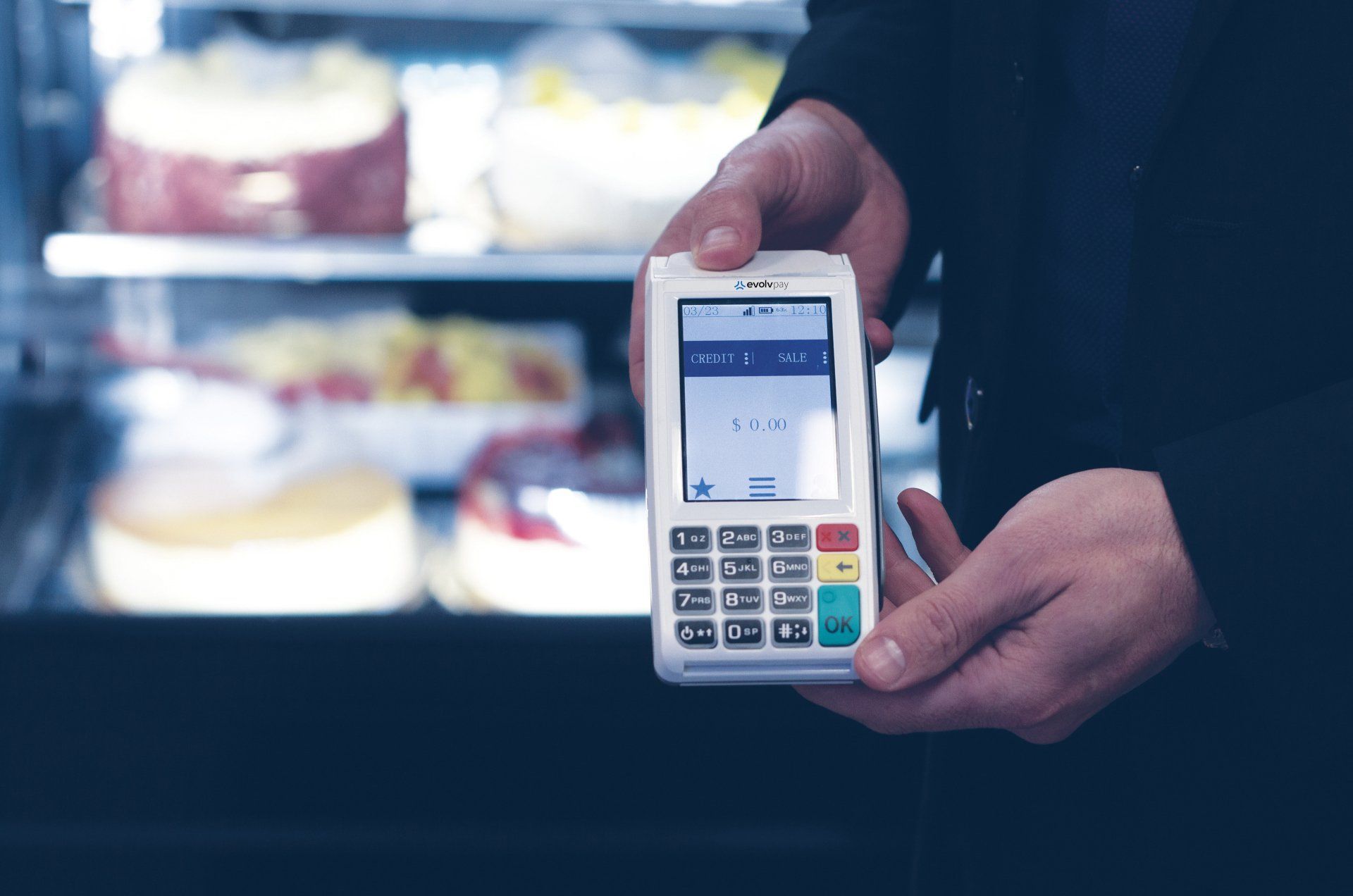A person is holding a credit card machine in front of a display of cakes.