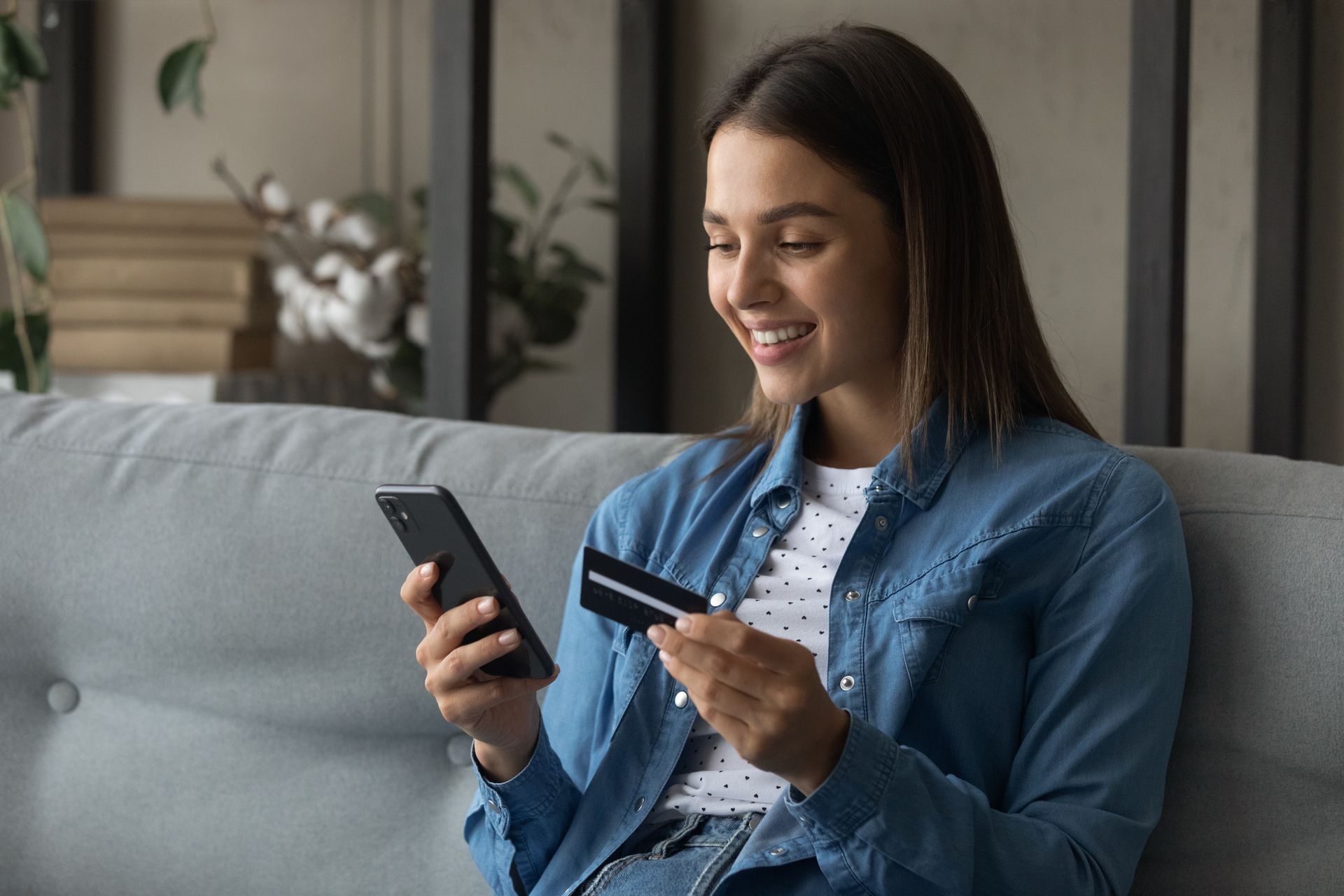 A woman is sitting on a couch holding a credit card and a cell phone.