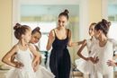 Ballet instructor in a black dress guiding four young dancers in white tutus in a studio