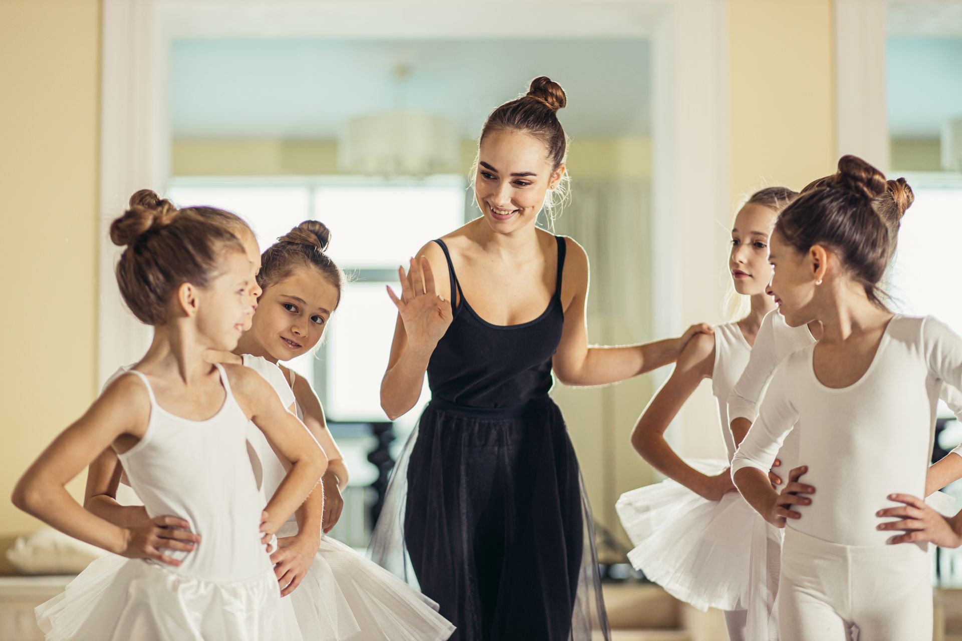 Ballet instructor in a black dress guiding four young dancers in white tutus in a studio