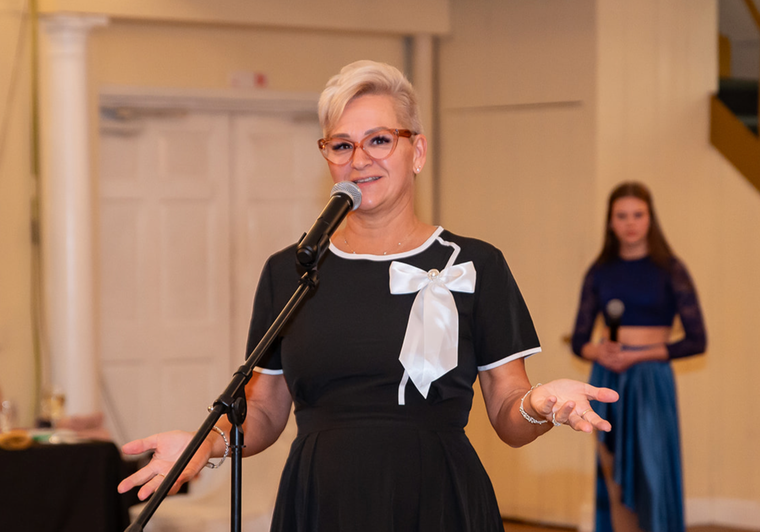 Woman speaking at a microphone in a black dress with a white bow, indoors, with another woman in the background.