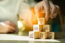 Hand placing glowing block on stacked cubes with business icons, in a soft-lit workspace