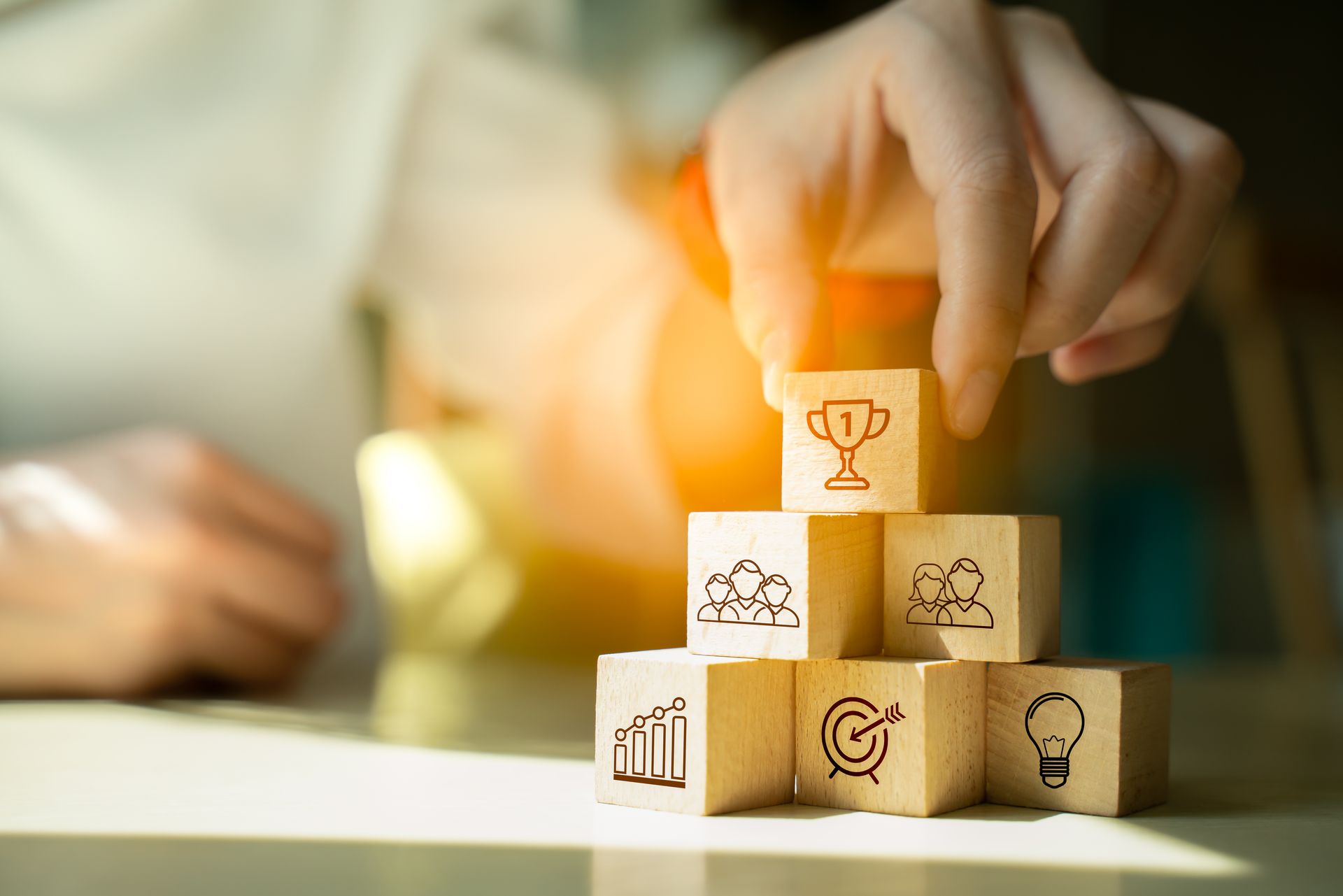 Hand placing glowing block on stacked cubes with business icons, in a soft-lit workspace