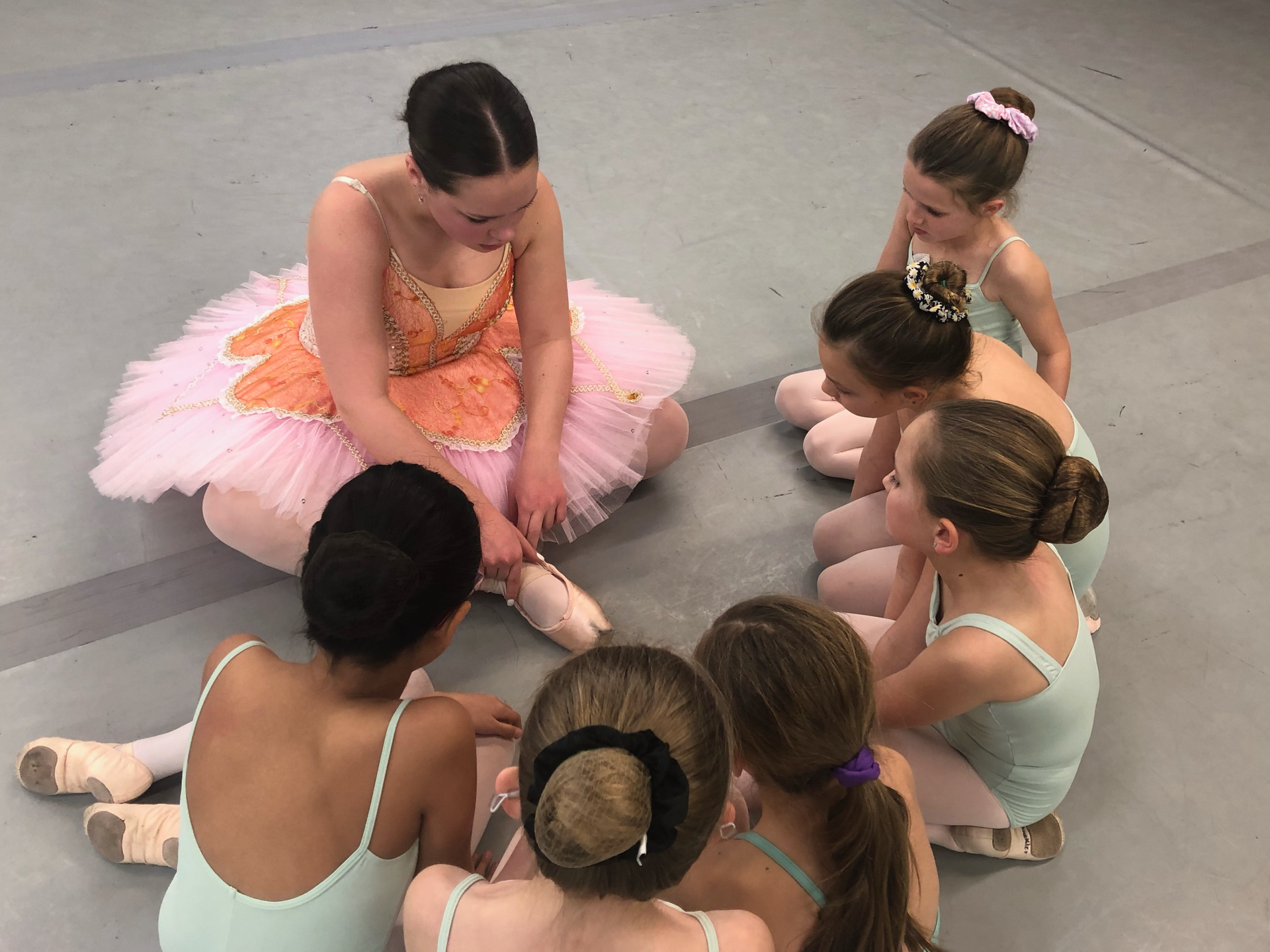 Children in ballet tutus sitting in a circle on a studio floor, practicing together
