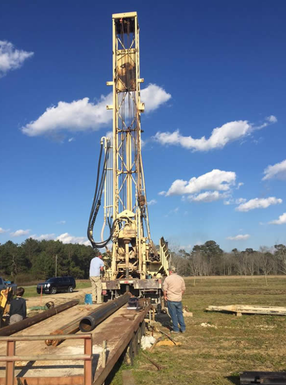 Well drilling rig in a field with two men standing nearby. Blue sky with clouds.