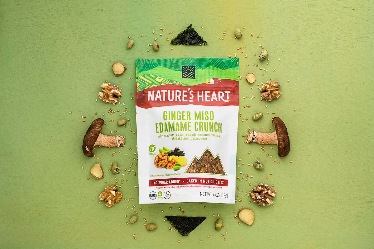 An assortment of dried mushrooms, walnuts, edamame, ginger and seaweed on a green background, around Nature's Heart Ginger Miso Edamame Crunch snack mix. Product photography for Nature's Heart by JAM Creative.