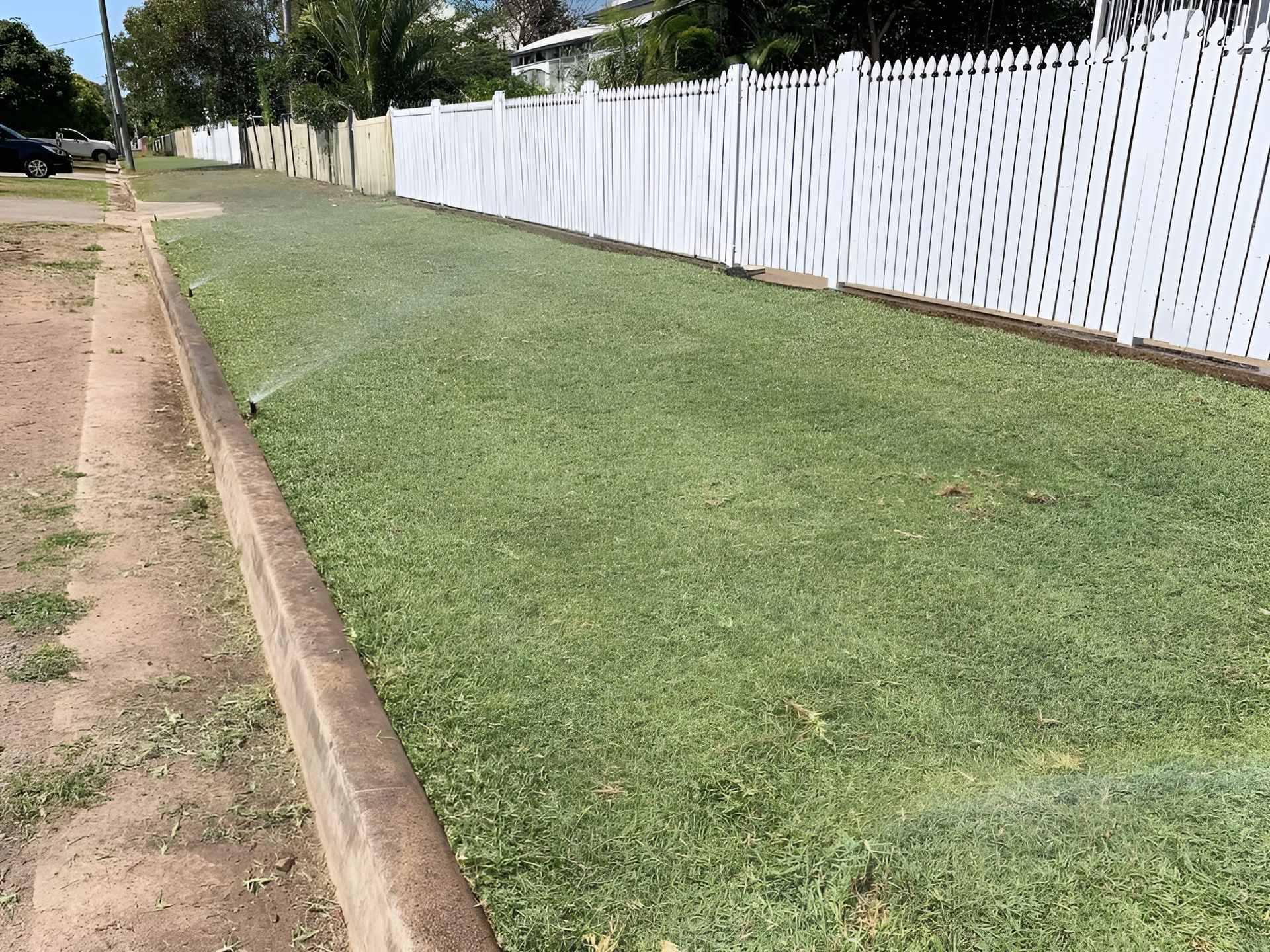 Lawn with Sprinklers Running Next to A White Picket Fence and A Concrete Curb — Top Notch Turf in Mount Isa, QLD
