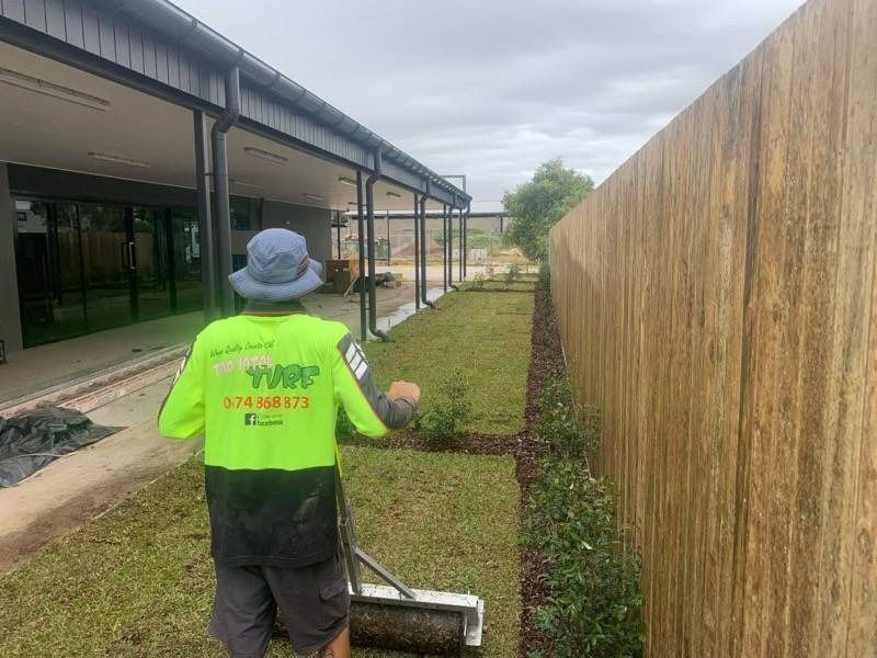 Man Rolling Lawn, Wearing a Hi-Vis Shirt, Next to A Wooden Fence and Building — Top Notch Turf in Rasmussen, QLD