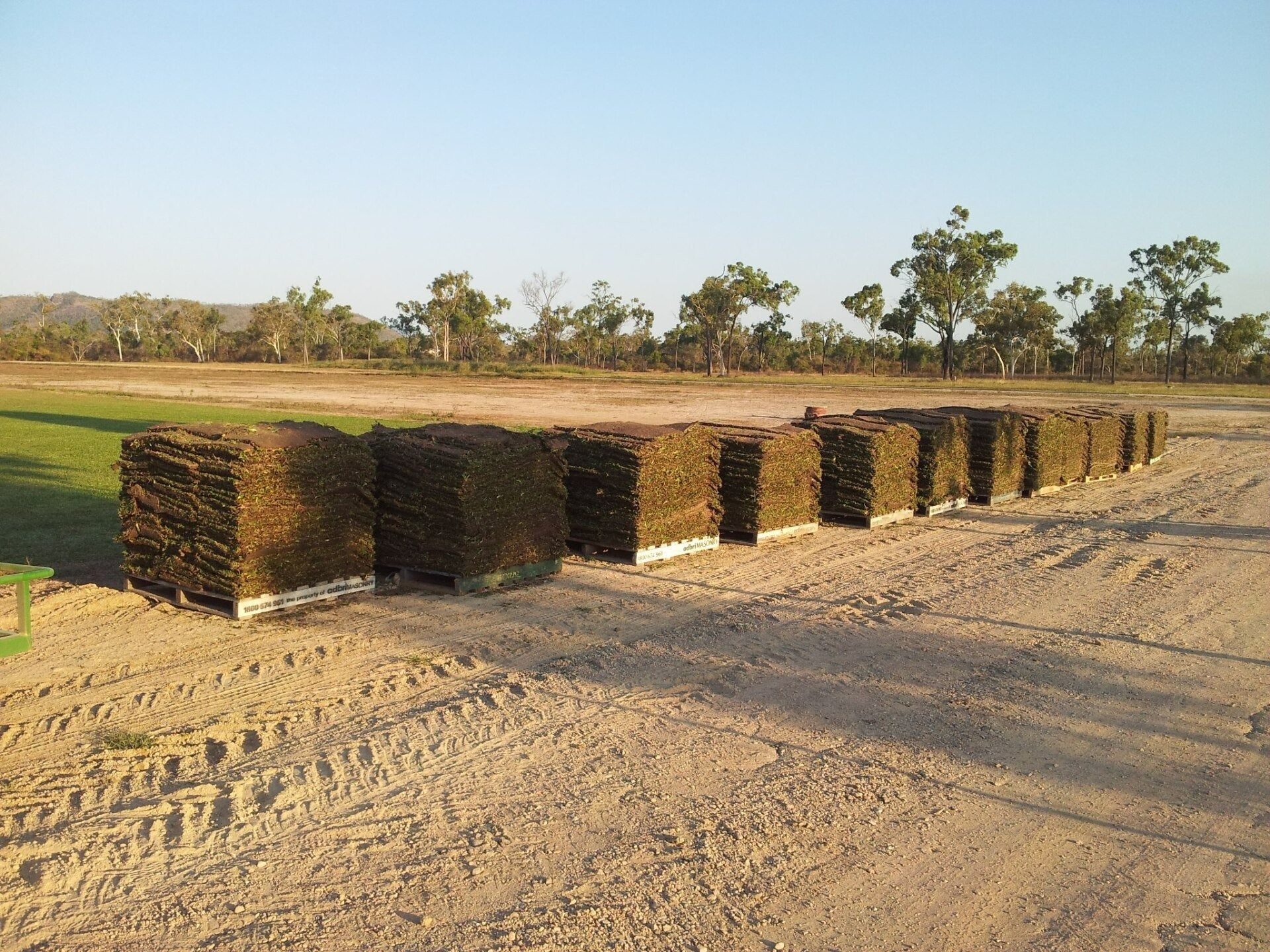 Pallets of Cut Sod Arranged on Dirt Field, Trees and Hills in Background Under Blue Sky — Top Notch Turf in Rasmussen, QLD