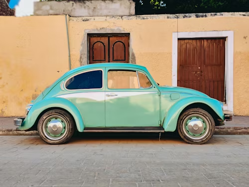 Teal vintage Volkswagen Beetle parked on a cobblestone street in front of a yellow building with brown doors.