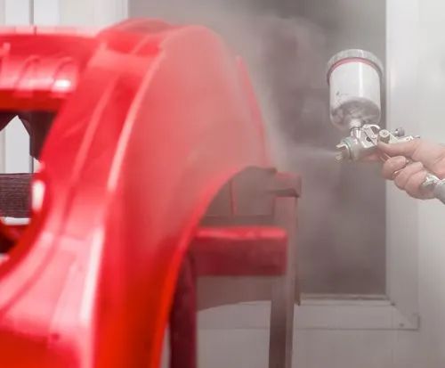 Spraying red metal part with paint gun, creating a fine mist in a workshop