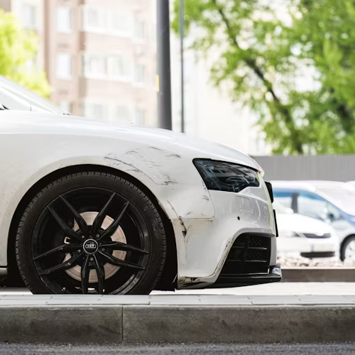 White car with damage to the front quarter panel and a black rim, parked outside, daylight.