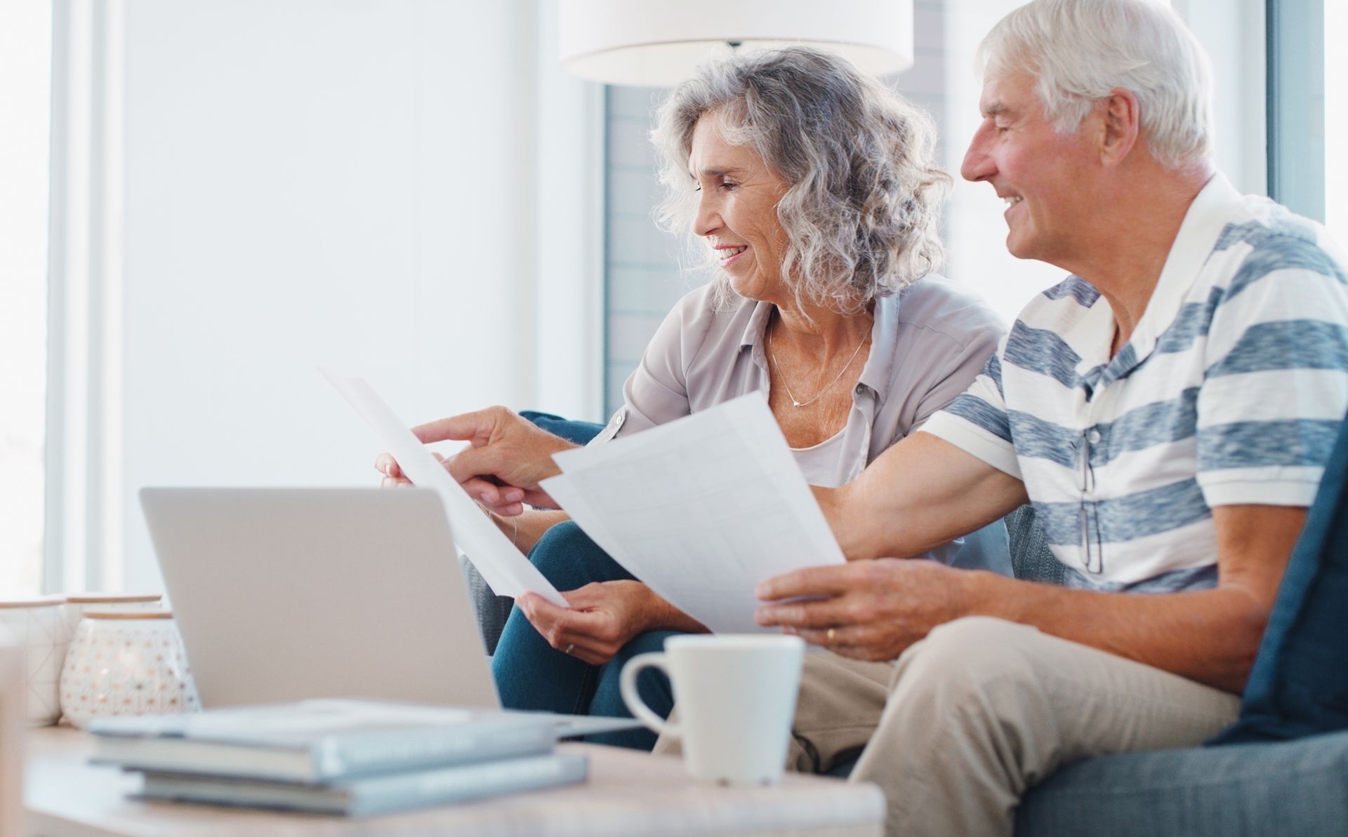 Older couple reviewing documents with a laptop and coffee mug on a couch.
