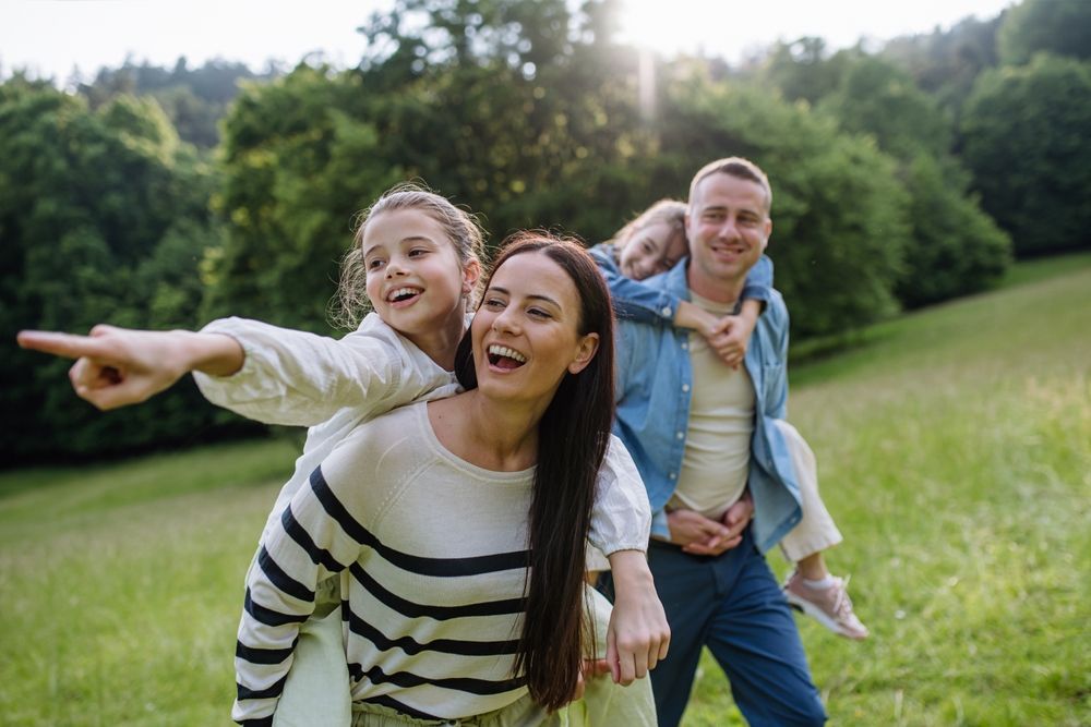 Family of four smiling and laughing outdoors; sunny day, piggyback rides in a grassy field.